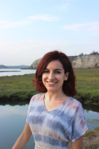 Photo of Alexa Tierno for LEAD. Alexa has red hair and wears a blue striped shirt while posing in front of a marsh with water, grasslands, and cliffs.