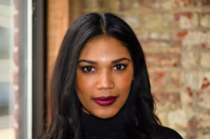 Headshot photo of Alysia Cruz for LEAD. Alysia wears a black shirt and burgandy lipstick while posing in front of a rustic brick wall