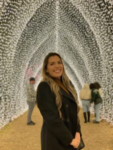 Photo of Barbara Martinez Neda for LEAD. Barbara has long blonde hair and a black winter jacket while posing in front of a tunnel of white holiday lights