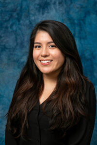 Photo of Celina Morales for Lead. Celina has long dark hair and a black blazer while posing in front of a tie-dye blue background