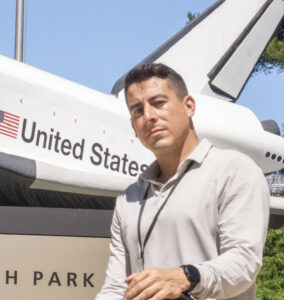 Photo of Cody Gonzalez for LEAD. Cody has short, dark hair and wears a white polo sweater while posing in front of an air plane with an American Flag and large text that says "United States"