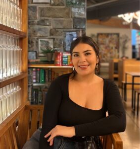 Photo of Julybeth Murillo for LEAD. Julybeth wears a black shirt and sits in in a wood chair in front of a stone wall with books and glass bottles