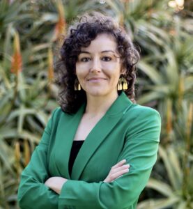 Photo of Jeanie Toscano for LEAD. Jeanie wears a green blazer and gold earrings while posing in front of a flowering plant