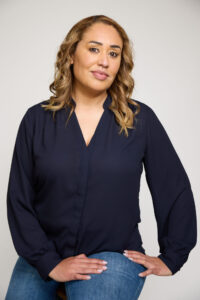 Photo of Veroncia Gonzalez for LEAD. Veronica wears a dark blue blouse and blue jeans while posing in front of a plain gray background