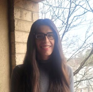 Photo of Victoria Knapp Perez. Victoria poses with long brown hair and a gray turtleneck shirt infront of a tree and brick wall