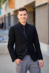 Photo of Zachary Velazquez for LEAD. Zachary wears a black button up with a black tie and jacket, and gray pants. The background is an outdoor hallway with columns